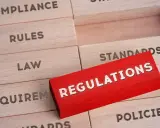 Drewniane klocki z napisami: "Compliance", "Rules", "Law", "Requirements", "Standards", "Policies". Czerwony klocek z napisem "REGULATIONS" wyróżnia się na tle pozostałych, symbolizując kluczowe środki trwałe w biznesie.