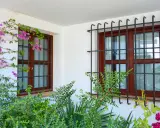 Dos ventanas con barrotillo de hierro y marcos de madera, rodeadas de vegetación y flores moradas.