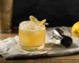 En gyllene whiskey drink med is och citronskal, redo att avnjutas. Bredvid står en jigger och en citron.