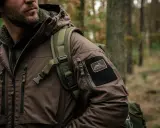 Mężczyzna w brązowej kurtce z naszywką Helikon-Tex, symbol polskie marki sportowe, w lesie.