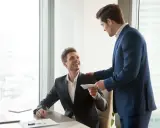 Szef wręcza pracownikowi kopertę z premią uznaniową. Uśmiechnięty mężczyzna siedzi przy biurku, otrzymując zasłużone docenienie.