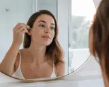 Une femme applique un sérum à l'acide glycolique sur son visage, reflétée dans un miroir.