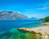 Ein idyllischer Gardasee Strand mit kristallklarem Wasser, felsigen Ufern und majestätischen Bergen im Hintergrund.