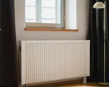 Biały grzejnik pod oknem, obok ciemnej zasłony i lampy stojącej. Dowiedz się, jak ustawić grzejniki przy ogrzewaniu gazowym dla optymalnego komfortu.