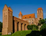 Potężny zamek w Kwidzynie z cegły, z charakterystycznymi wieżami i arkadowym murem obronnym, lśni w słońcu na tle błękitnego nieba.