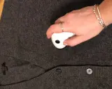 En hand med smycken använder en noppmaskin på en mörkgrå kofta. Materialet känns mjukt, kanske rayon material svenska.