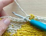Mano tejiendo con ganchillo, creando un punto bajo con hilo blanco sobre una tela amarilla y blanca.