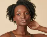 Une femme à la peau foncée, aux cheveux afro volumineux et bouclés, porte des boucles d'oreilles dorées.