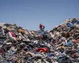 Un uomo in maglia rossa cammina su una montagna di vestiti scartati, simbolo dell'inquinamento tessile.