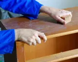 Hände schleifen eine Holzoberfläche mit einem Schleifklotz. So lernt man, wie lackiere ich richtig für ein glattes Ergebnis.
