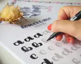 Mano practicando cómo hacer lettering con un rotulador sobre un papel con letras de ejemplo.
