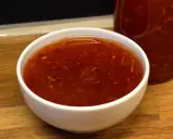 En skål med glansig, röd sweet chili sås med små bitar av chili och vitlök. En flaska med samma sås syns i bakgrunden.