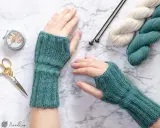 Manos con guantes tejidos a mano, listos para un nuevo patron. Lana, agujas y tijeras decorativas completan la escena.