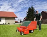 Mężczyzna w zielonym stroju pcha wertykulator, kiedy wertykulujemy trawnik. W tle dom i biała furgonetka.
