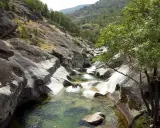 Un río de aguas cristalinas fluye entre rocas erosionadas, formando piscinas naturales. Dos personas disfrutan del baño en uno de los pilones, rodeados de vegetación y montañas.
