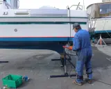 Un homme en combinaison bleue travaille sur la coque d'un bateau pour enlever l'antifouling. Des outils et des pots de peinture sont à proximité.