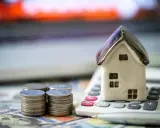 Miniaturowy domek na kalkulatorze, stosy monet i banknoty. Czy zadatek jest zwrotny? Finansowe rozważania o nieruchomościach.
