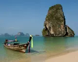En traditionell longtailbåt flyter i turkost vatten vid en sandstrand i Thailand. Klippöar reser sig ur havet. Kanske en resa hit inkluderar vaccin Thailand pris.