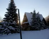 Zimowy krajobraz z bacówką pod Małą Rawką. Śnieg sypie, a słońce przebija się przez drzewa.