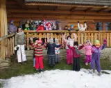 Wesoła gromadka dzieci pozdrawia z bacówki u harnasia. Wszyscy ubrani w zimowe stroje, radośnie machają rękami na tle drewnianego domu.