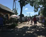 Ludzie spacerują po targu, podziwiając **tleń atrakcje**. Widać stoiska z towarami, restaurację i drzewa rzucające cień.