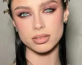 Ojos ahumados en tonos rosados y marrones, realzando unos ojos azules penetrantes. Labios nude y cabello oscuro completan el look.