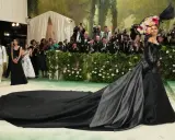 Una mujer con un vestido negro y un tocado floral posa para las cámaras en la alfombra verde, mientras un hombre ajusta la cola de su vestido, todo bajo el tema de la Met Gala.