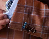 A man adjusts his watch on his wrist, revealing the detailed pick stitching on the cuff of his brown plaid suit jacket.