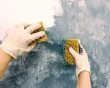 Händer med handskar målar akryl med svamp på en vägg för att skapa en texturerad effekt.