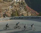 Ciclisti affrontano la salita del Col de la Couillole, con un villaggio arroccato sulla montagna autunnale.
