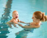 Radość dziecka podczas pierwszego razu na basenie. Mama trzyma malucha w wodzie, oboje uśmiechnięci.