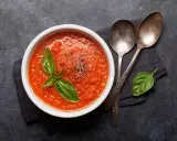 Schüssel mit selbstgemachter Tomatensauce, garniert mit Basilikum und Pfeffer, daneben zwei Löffel auf dunklem Hintergrund.