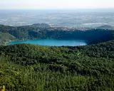 Vista panoramica del lago di Albano, circondato da rigogliosa vegetazione, con il borgo di Castel Gandolfo e Rocca di Papa in lontananza.