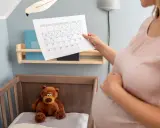Ciężarna kobieta sprawdza kalendarz, zaznaczając dni do terminu porodu. W tle widać łóżeczko z misiem.