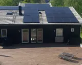 Ett mörkt hus med en liten solcellsanläggning på taket. En stor altan med solstolar leder ut mot en grönskande trädgård.