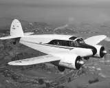 Ein zweimotoriges Flugzeug, ein klassisches Cessna-Modell, fliegt über eine ländliche Landschaft mit einer Stadt und Industrieanlagen im Hintergrund.
