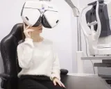 Kobieta w białym swetrze siedzi na czarnym fotelu, badana przez optometrystę na nowoczesnym sprzęcie.