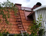 Takstege på ett rött tegeltak, en del av taket syns genom lövverk. Kanske för att följa takstege regler vid sotning.