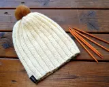 En ribbstickad mössa med en brun pompom ligger på ett träbord bredvid stickor. Perfekt för ett ribbstickad mössa gratis mönster.