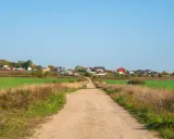 Droga gruntowa prowadzi do osiedla domów. Minimalna szerokość drogi dojazdowej do działki budowlanej jest tu widoczna, otoczona polami i trawami.