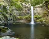 La fervenza do Pombar, una cascada serena rodeada de rocas cubiertas de musgo y vegetación exuberante, cae en una poza de aguas tranquilas.