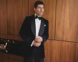 A man in a dark velvet tuxedo with a bow tie, embodying festive chic. He stands by a modern fireplace with flames.
