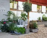 En lummig kvarnkulla trädgård med blommor i krukor och rabatter. En vit bänk och dekorativa spaljéer skapar en charmig miljö.