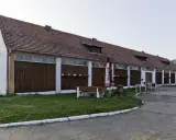 Długi budynek stajni z drewnianymi drzwiami i oknami, z parasolem i ławką przed nim.