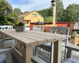 Bygga utebord: En person mäter trä för att bygga ett robust utomhusbord. Ett färdigt bord står på en altan med blommor och hus i bakgrunden.