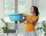 Une femme tente de contenir une fuite d'eau avec un bac bleu, visiblement exaspérée par le dégât des eaux voisin récalcitrant.