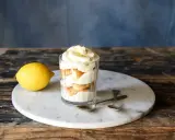 Ein Glas mit geschichtetem Zitronen Tiramisu, garniert mit Zitronenschale. Daneben liegt eine Zitrone.