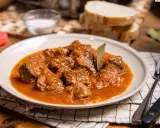 Sabrosa carne con tomate, guisada lentamente hasta quedar tierna, servida en un plato blanco con pan y sal al lado.