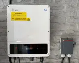 En vit GoodWe växelriktare 10kW monterad på en grå stenvägg. Bredvid sitter en grå ABB-box.
