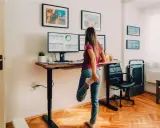 Femme s'étirant près de son bureau réglable en hauteur, pour un confort au bureau optimal. Deux écrans affichent des graphiques.